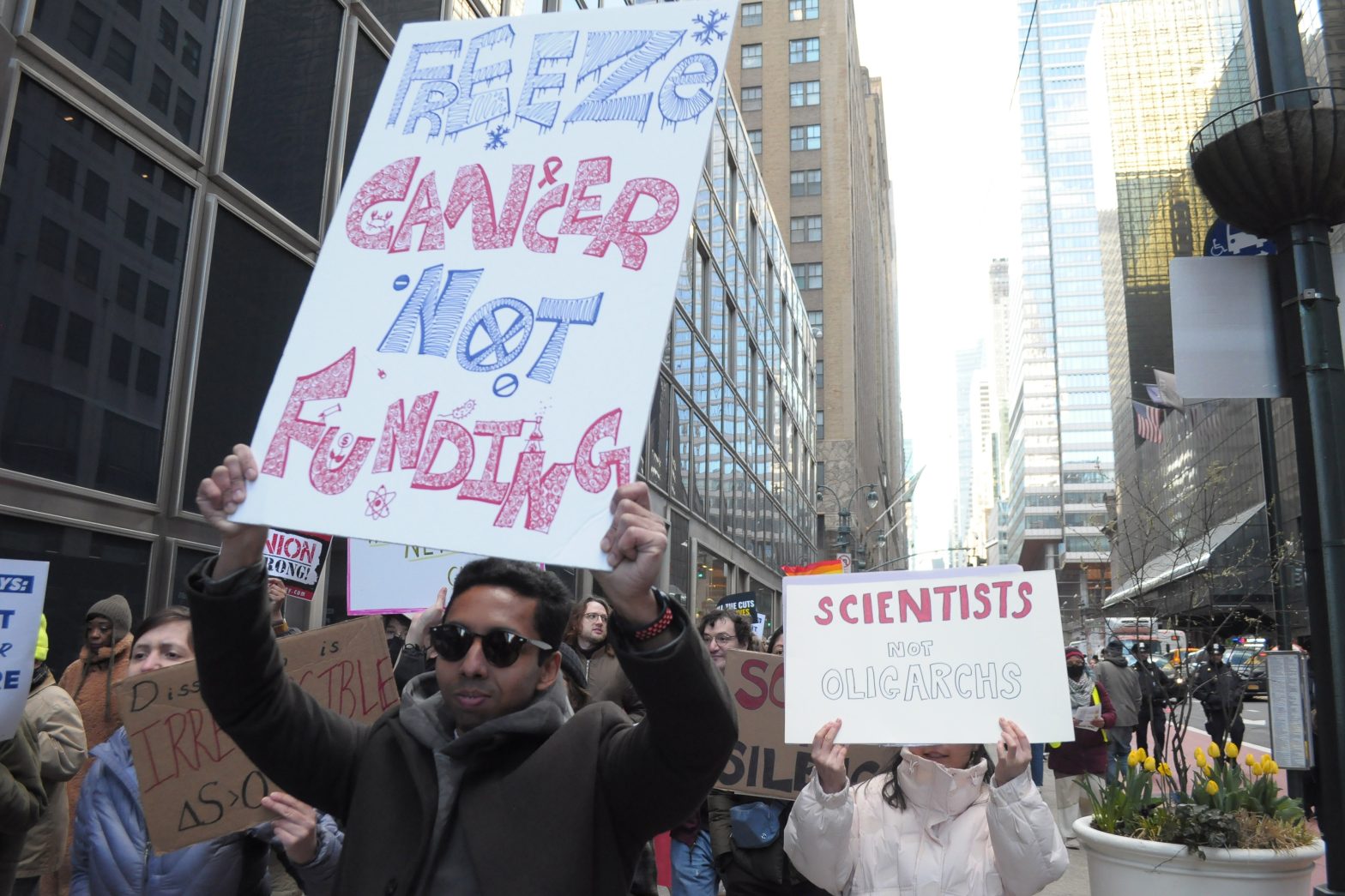 Demonstrators march in a street holding signs, including one that says "Freeze cancer, not funding"